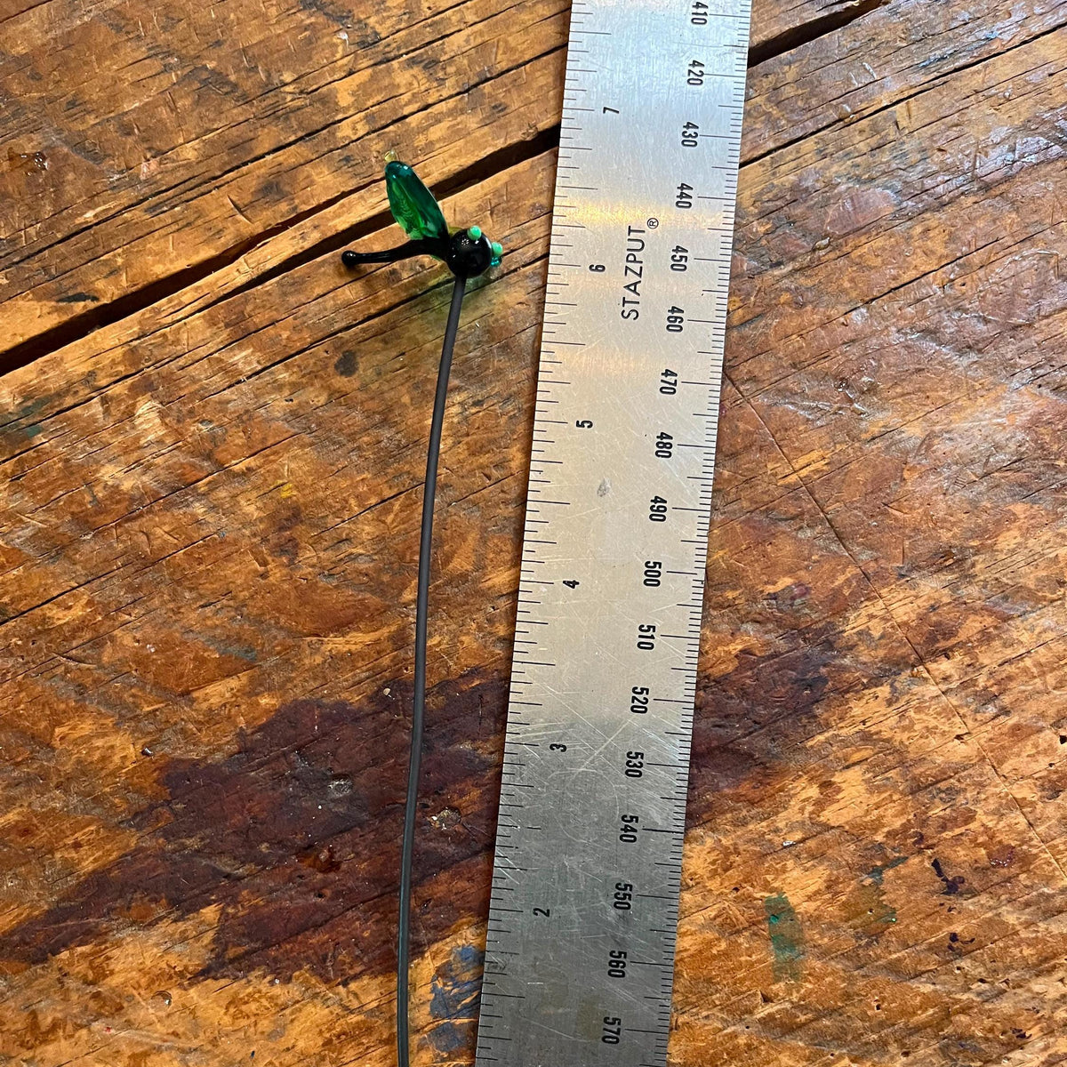 A long, thin black tool with a small green and black end piece is placed next to a metal ruler on a worn wooden tabletop.