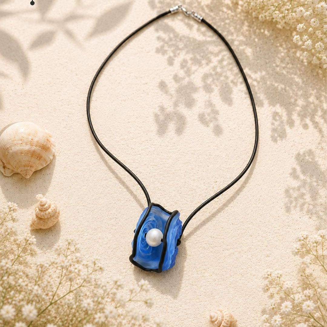 A blue and black glass pendant necklace with a white pearl accent, displayed on a sandy background with seashells and dried flowers. The brand logo 'Single Splash' is visible in the top left corner.