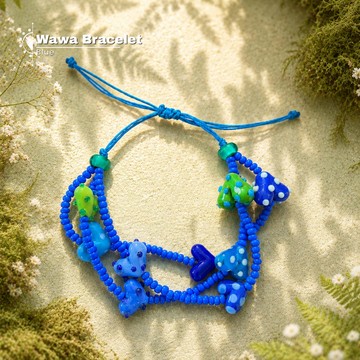 A blue beaded bracelet with fish and sea creature charms, displayed on a sandy surface with green moss and dried flowers. The brand name 'Wawa Bracelet Blue' is visible in the top left corner.