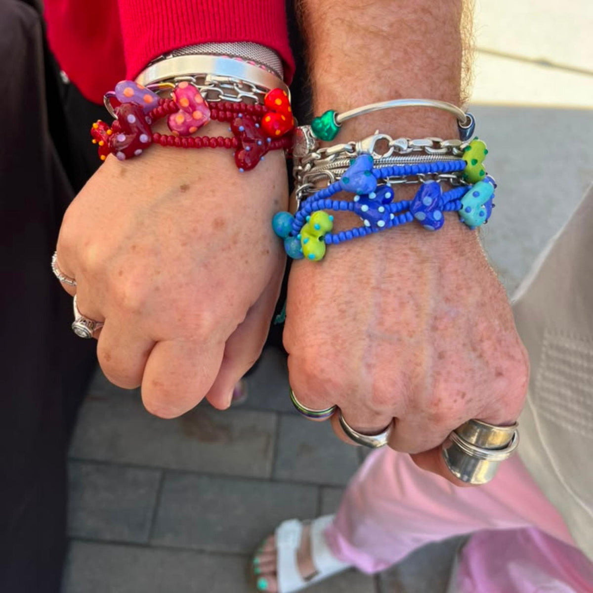Two people standing close together, each wearing multiple colorful beaded bracelets on their wrists. The bracelets feature red, blue, pink, green, and white beads, with one bracelet showing a silver-toned skull charm and another showing a blue skull charm.