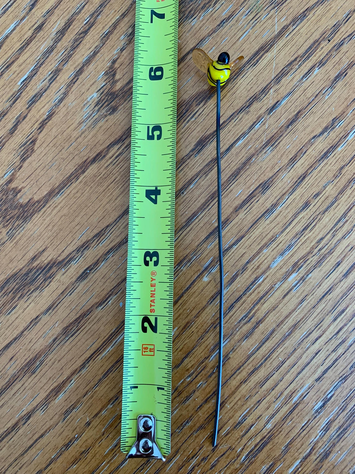 A small yellow and black bee-shaped object with a long thin black stem lies on a wooden surface next to a green Stanley tape measure for scale.