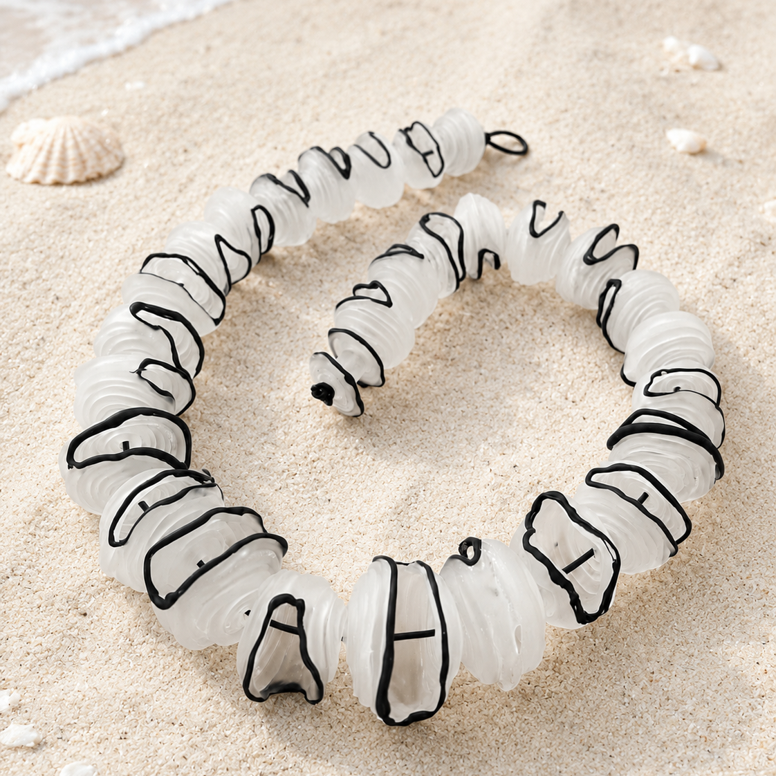 A coiled white rope or cord with black stitched or outlined accents, photographed on sandy ground with small shells and a bit of water visible in the corner.