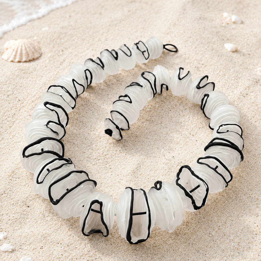 A coiled white rope or cord with black stitched or outlined accents, photographed on sandy ground with small shells and a bit of water visible in the corner.