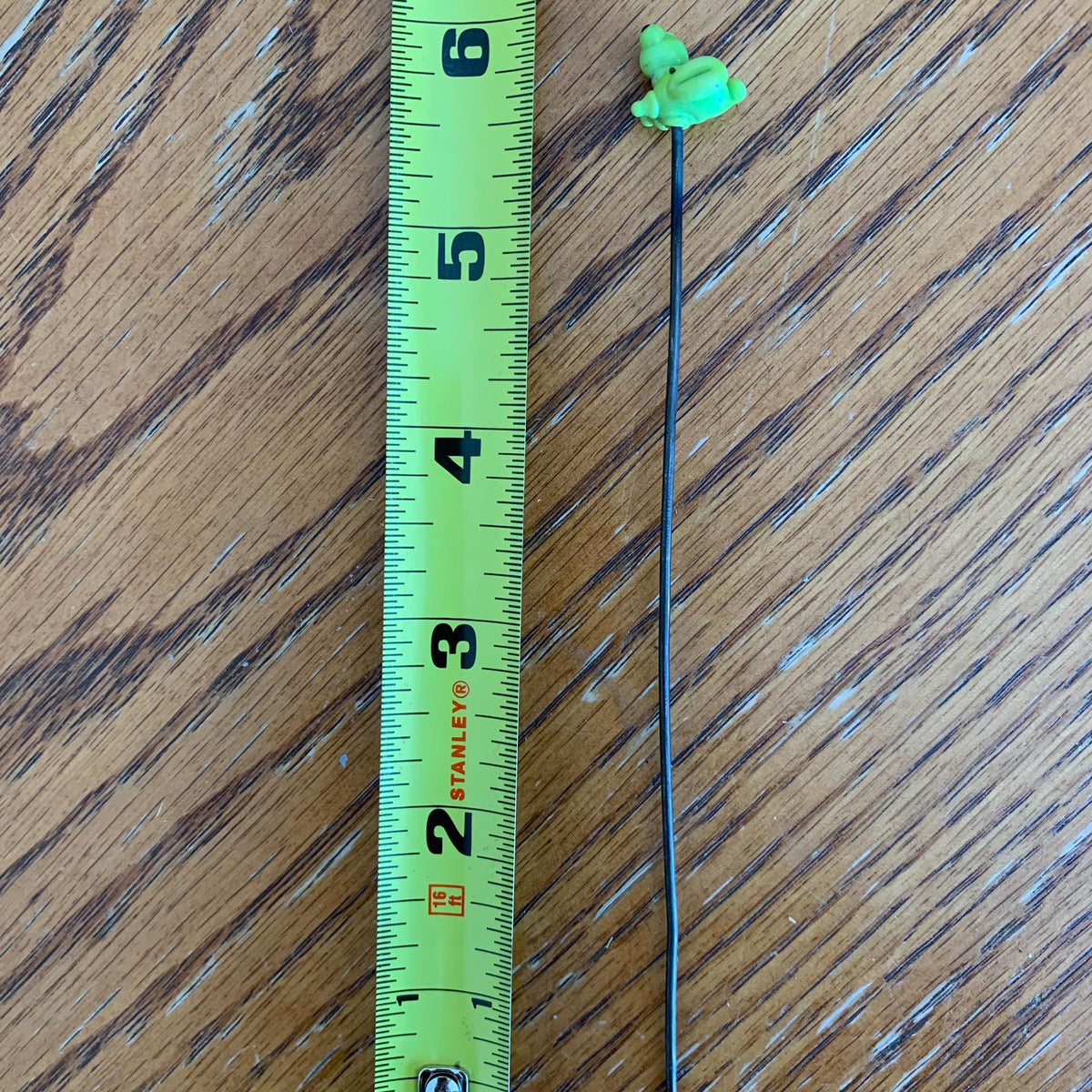 A small green frog-shaped hair pin on a black wire, shown next to a yellow Stanley tape measure on a wooden table.