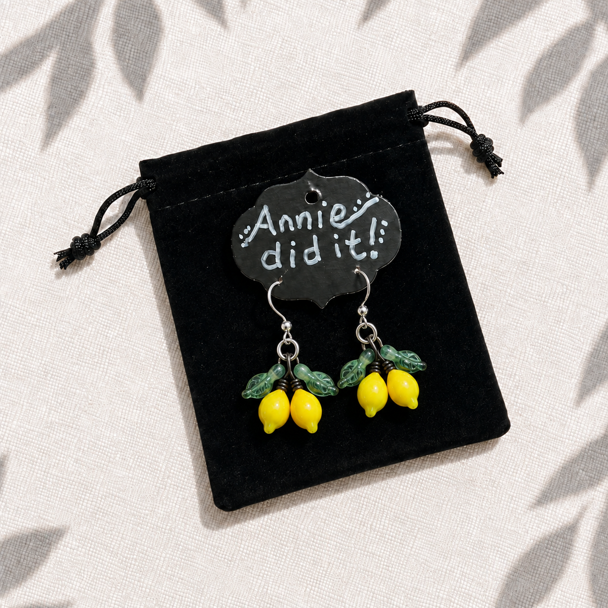 A pair of dangling earrings shaped like yellow lemons with green leaves, displayed on a small black velvet pouch that has a chalkboard-style tag reading 'Annie did it!!'.