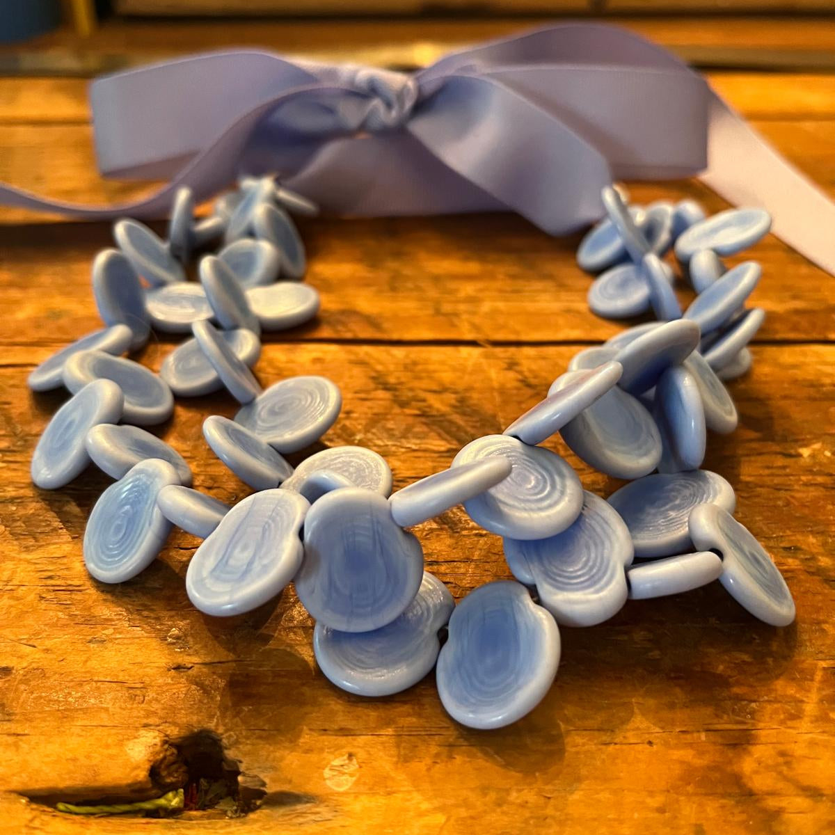 A light blue layered disc necklace with a matching light blue ribbon bow, displayed on a rustic wooden surface.
