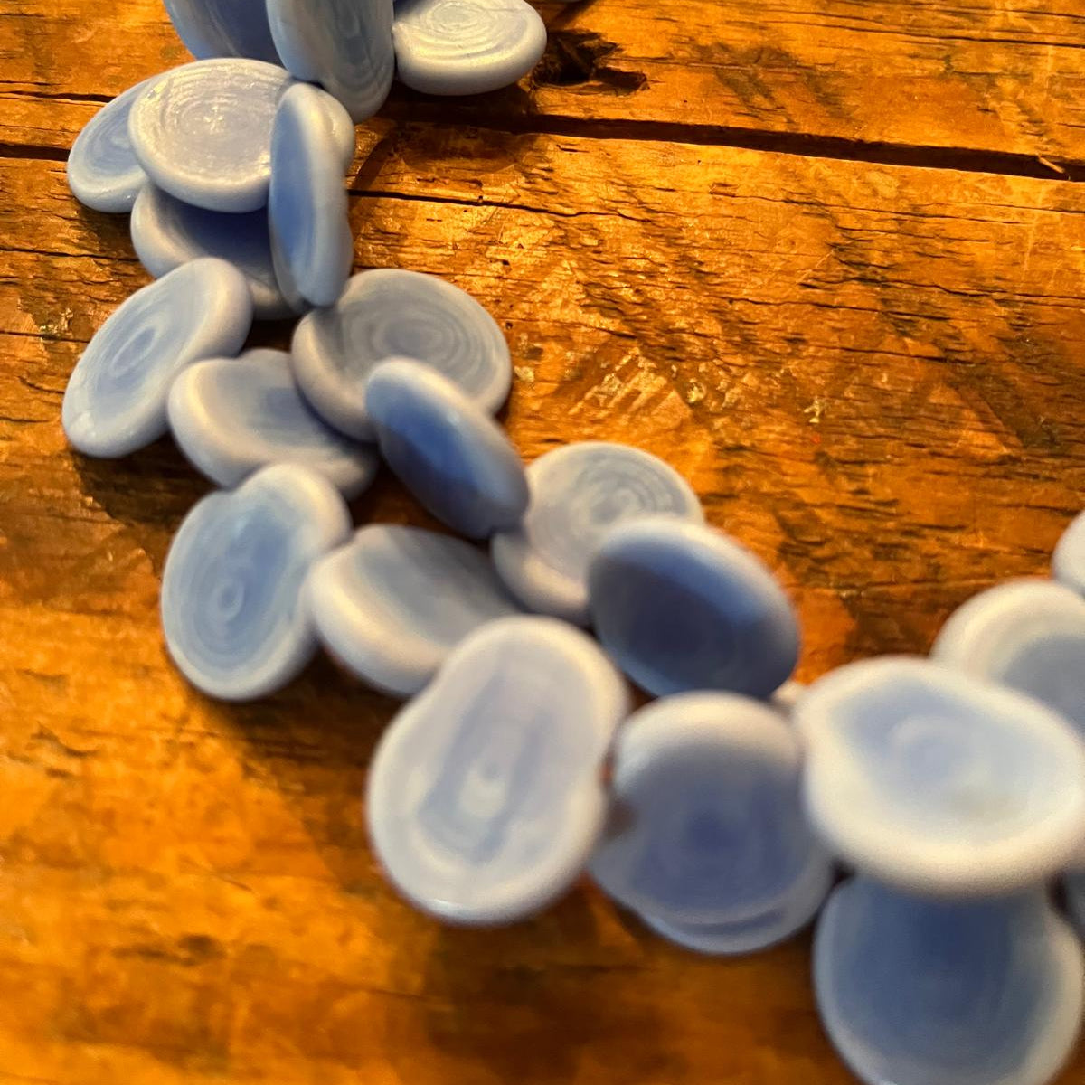 A close-up of a beaded necklace made of flat, oval-shaped light blue and white beads with concentric ring patterns, resting on a rustic wooden surface.