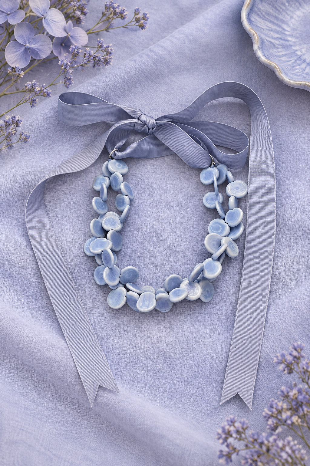 A handmade necklace made of light blue flower-shaped beads with a dusty lavender ribbon tied in a bow at the top, displayed on a rustic wooden surface.