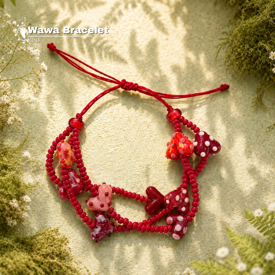 Red beaded bracelet with multiple charm beads, including a pink bear, red heart, and white polka dot motifs, displayed on a textured beige surface with green moss and small white flowers.