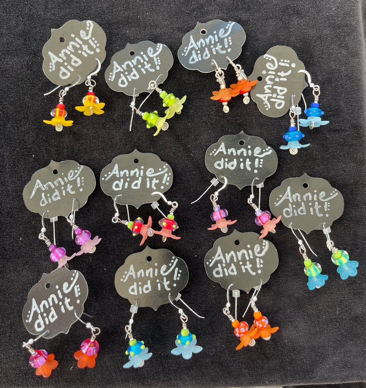 A set of colorful dangle earrings displayed on dark gray tags that read 'Annie did it!'. The earrings feature small, vibrant flower charms in yellow, orange, green, blue, pink, and light blue, suspended from silver hooks.