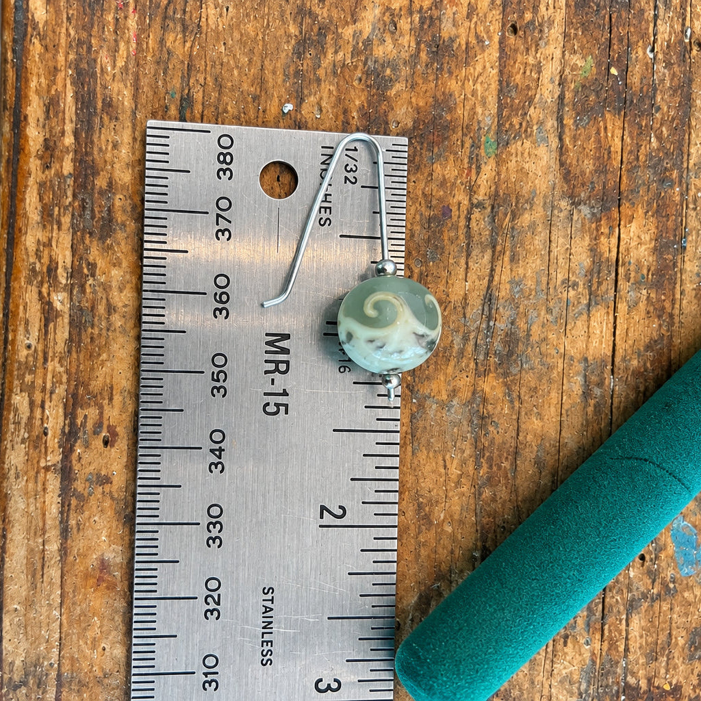 A small round marbled bead in white, gray, and green sits on a metal ruler on a wooden table, with a teal felt pencil or nail file above it and a person's feet visible at the bottom edge.