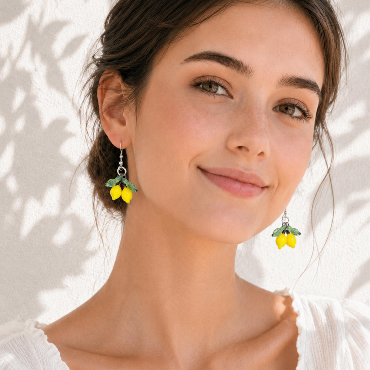 A woman wearing dangling lemon earrings, shown in a close-up portrait against a softly lit white wall with leaf shadows.