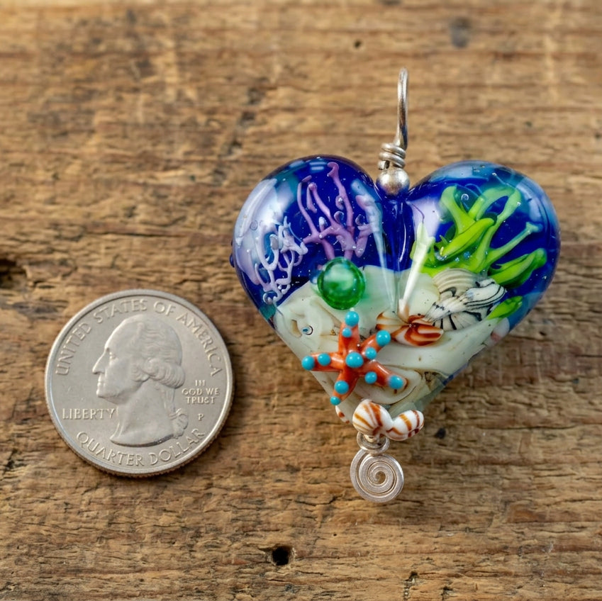 A heart-shaped glass pendant with a colorful coral reef design, shown next to a US quarter for scale on a plain white background.