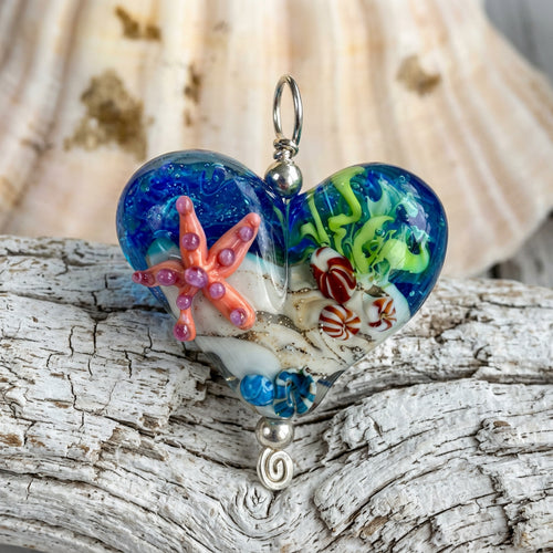A heart-shaped glass pendant charm with an ocean-themed design, featuring blue water, green sea plants, a pink starfish, and a red crab, displayed on a white seashell over a weathered gray wood surface.