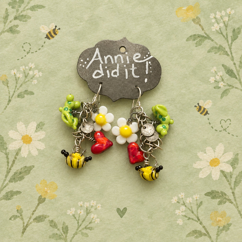 A handmade pair of dangling earrings displayed on a dark metal tag that reads 'Annie did it!' The earrings feature yellow and black bumblebees, white daisies with yellow centers, red hearts, and green floral charms on silver-toned hooks, all laid on a rustic wooden surface.