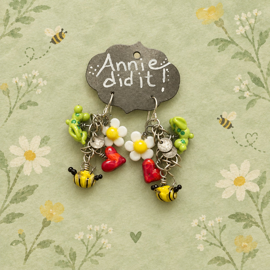 A handmade pair of dangling earrings displayed on a dark metal tag that reads 'Annie did it!' The earrings feature yellow and black bumblebees, white daisies with yellow centers, red hearts, and green floral charms on silver-toned hooks, all laid on a rustic wooden surface.