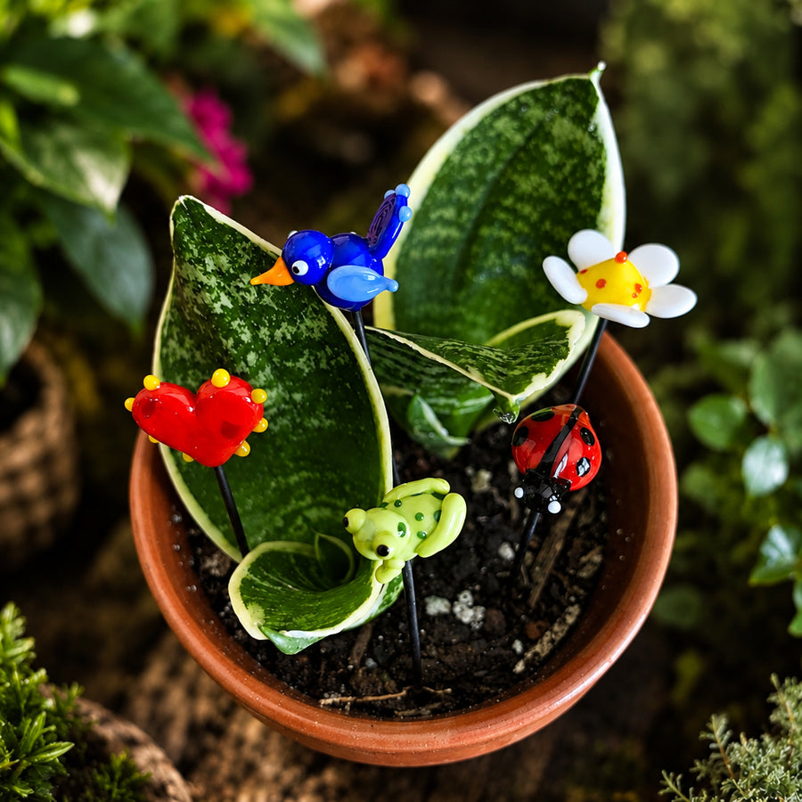 A terracotta pot containing a snake plant and four colorful glass garden stake decorations shaped like a blue bird, red heart, green frog, and ladybug, with a white and yellow flower. The background is a soft-focus garden setting with green foliage.