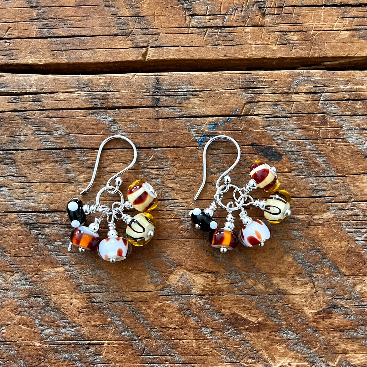 A pair of handmade dangle earrings with silver fishhook wires and clusters of colorful glass beads, including marbled orange, white, and yellow beads, resting on a rustic wooden surface.