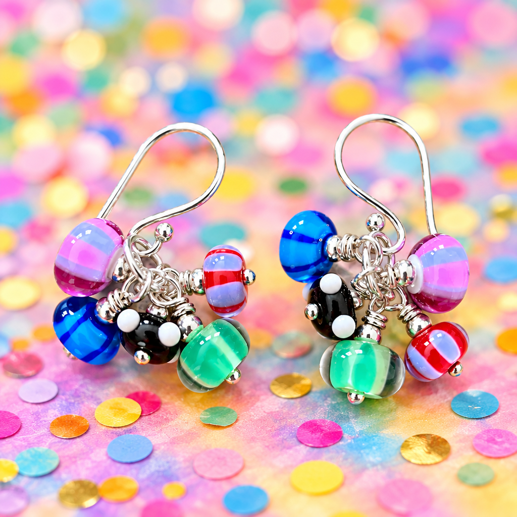 A pair of handmade glass bead drop earrings with silver fishhook wires, displayed on a rustic wooden surface. The earrings feature a cluster of colorful glass beads including blue, green, pink, black, red, and white, with one prominent pink and purple striped bead at the top of each earring.