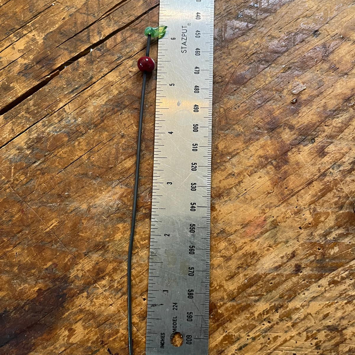 A long, thin black wire pin or hair stick with a small green leaf charm and a red bead, shown next to a metal ruler on a rustic wooden surface.