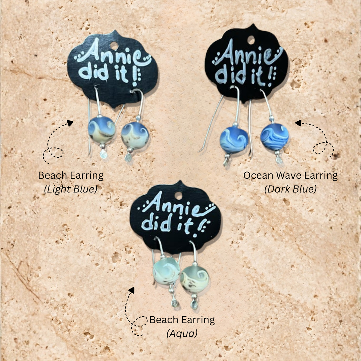 Three pairs of handmade dangle earrings displayed on black scalloped cards that read 'Annie did it!'. The cards are arranged on a sandy beige background, with text labels identifying the styles: 'Beach Earring (Light Blue)', 'Ocean Wave Earring (Dark Blue)', and 'Beach Earring (Aqua)'.