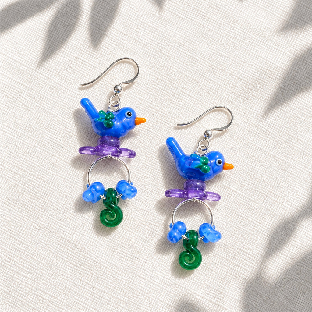 A pair of dangling earrings with blue bird charms, purple perched details, blue beads, and green spiral charms on silver hooks, displayed on a textured off-white fabric background.