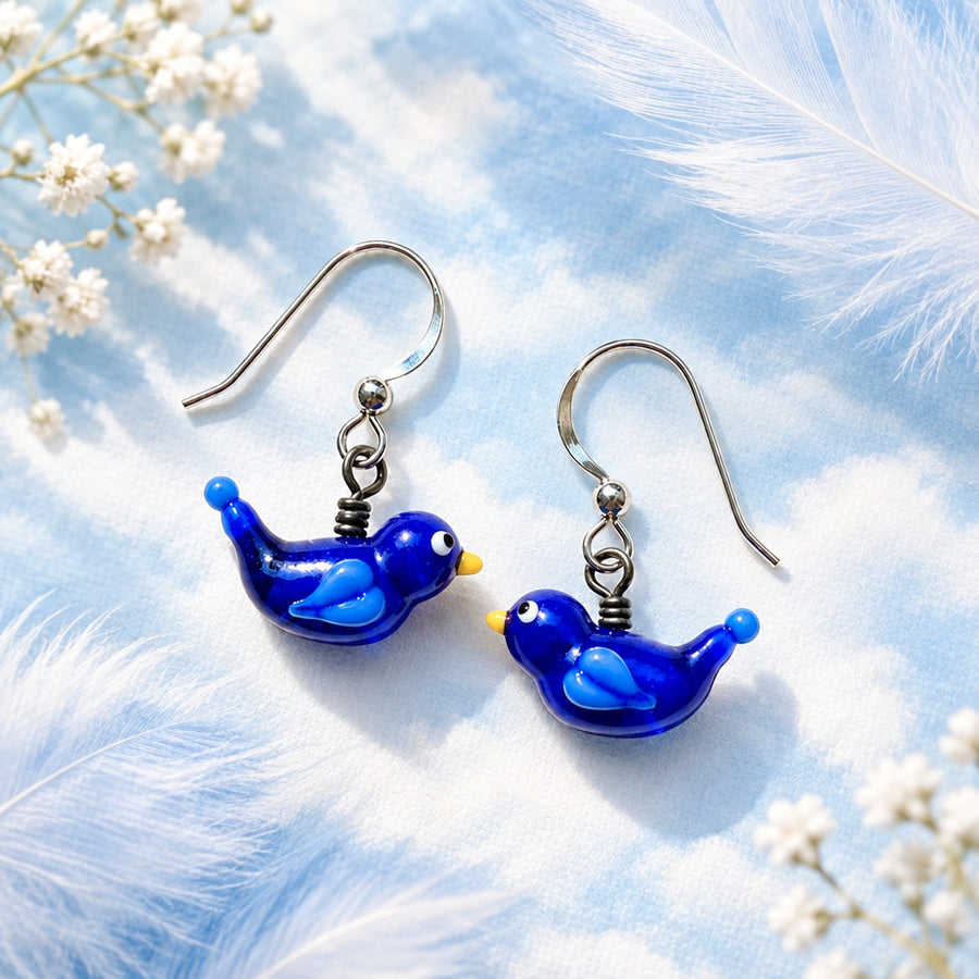 A pair of dangling bird earrings with glossy cobalt blue glass bodies, lighter blue wing details, and small yellow beaks, attached to silver fishhook wires. The earrings are displayed on a textured off-white fabric background with soft leaf shadows, and the word 'Earrings' appears in the top left corner.