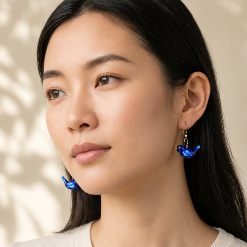 A woman wearing blue glass bird drop earrings, shown in a close-up portrait. The brand logo 'Earrings weekly 2' is visible in the top left corner.
