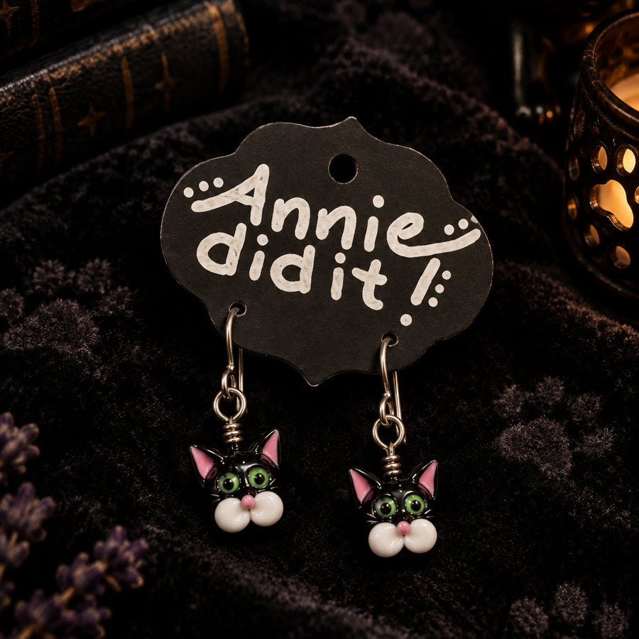 A pair of dangling earrings displayed on a black card that reads 'Annie did it!'. The earrings feature small cat faces with green and white details hanging from silver-toned hooks, set against a rustic wooden background.