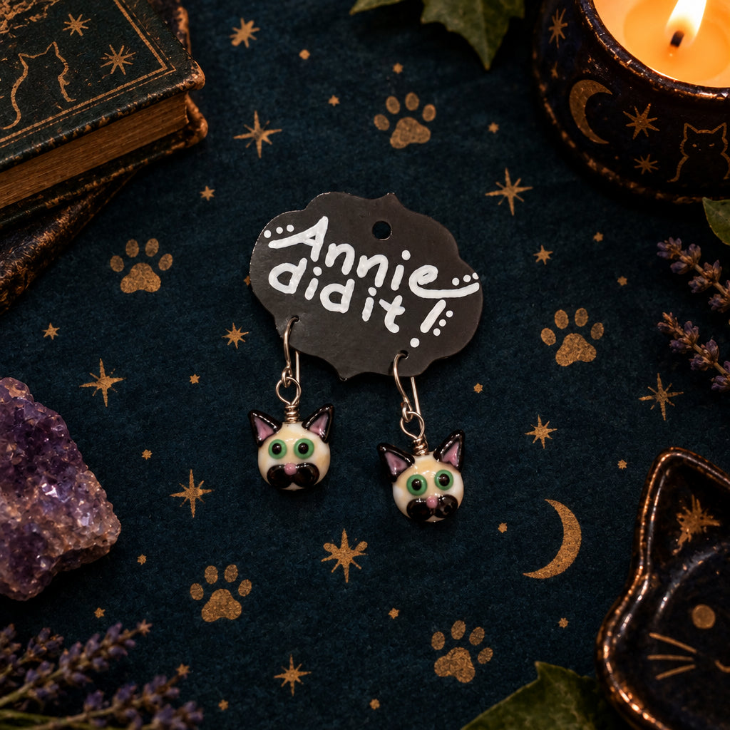 A pair of dangling earrings shaped like cat heads with green eyes and black ears, hanging from a black display card that reads 'Annie did it!' on a rustic wooden surface.