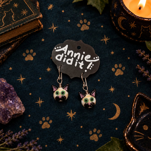 A pair of dangling earrings shaped like cat heads with green eyes and black ears, hanging from a black display card that reads 'Annie did it!' on a rustic wooden surface.