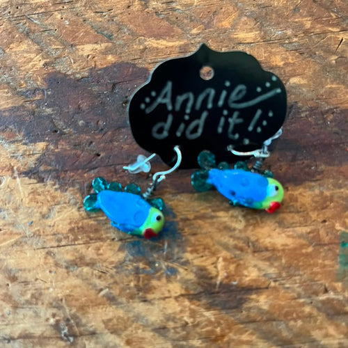 A pair of handmade parrot-shaped dangle earrings with blue, green, yellow, and red details, hanging from a black display card that reads 'Annie did it!' on a rustic wooden surface.