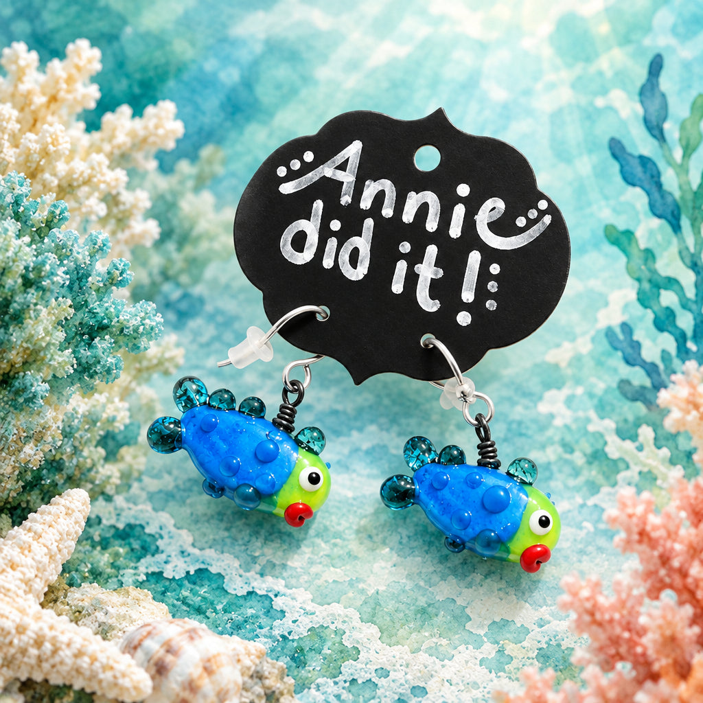 A pair of colorful fish-shaped dangle earrings displayed on a black cloud-shaped card that reads 'Annie did it!'. The earrings feature bright blue bodies, green heads, red noses, and small teal beads, shown on a textured white fabric background with soft leaf shadows.