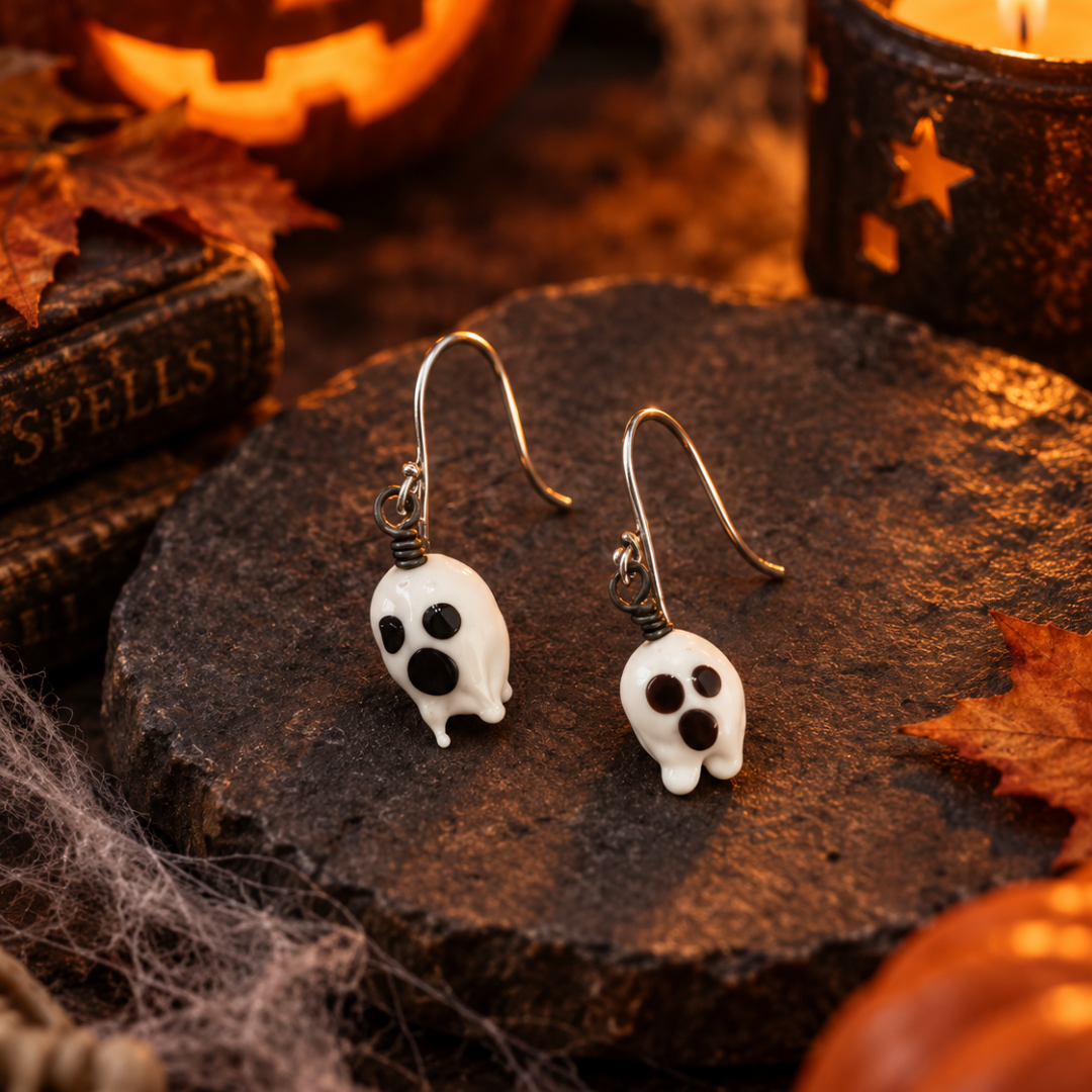 A pair of white skull-shaped dangle earrings with black facial features and silver hooks, displayed on a dark wooden slice surrounded by Halloween props including a lit candle, a carved pumpkin, autumn leaves, and an old book.