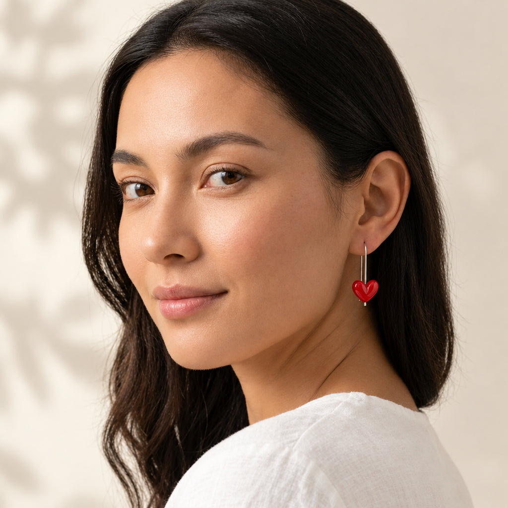 A woman wearing a single dangling heart-shaped red earring with a silver hook, shown in a close-up portrait against a soft beige background. The text 'Earrings' and 'eye Red' appear in the top-left corner.