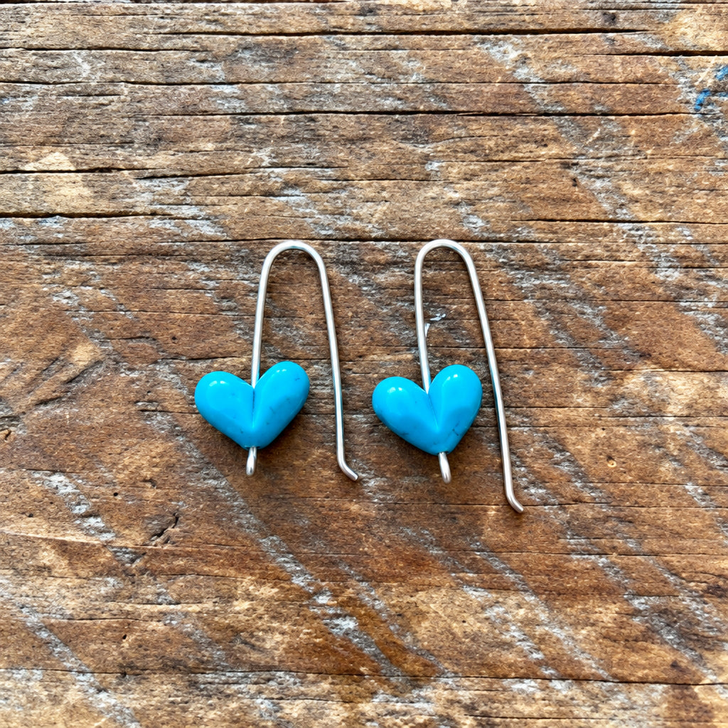 A pair of silver wire dangle earrings with small, bright blue heart-shaped beads, displayed on a rustic wooden surface.