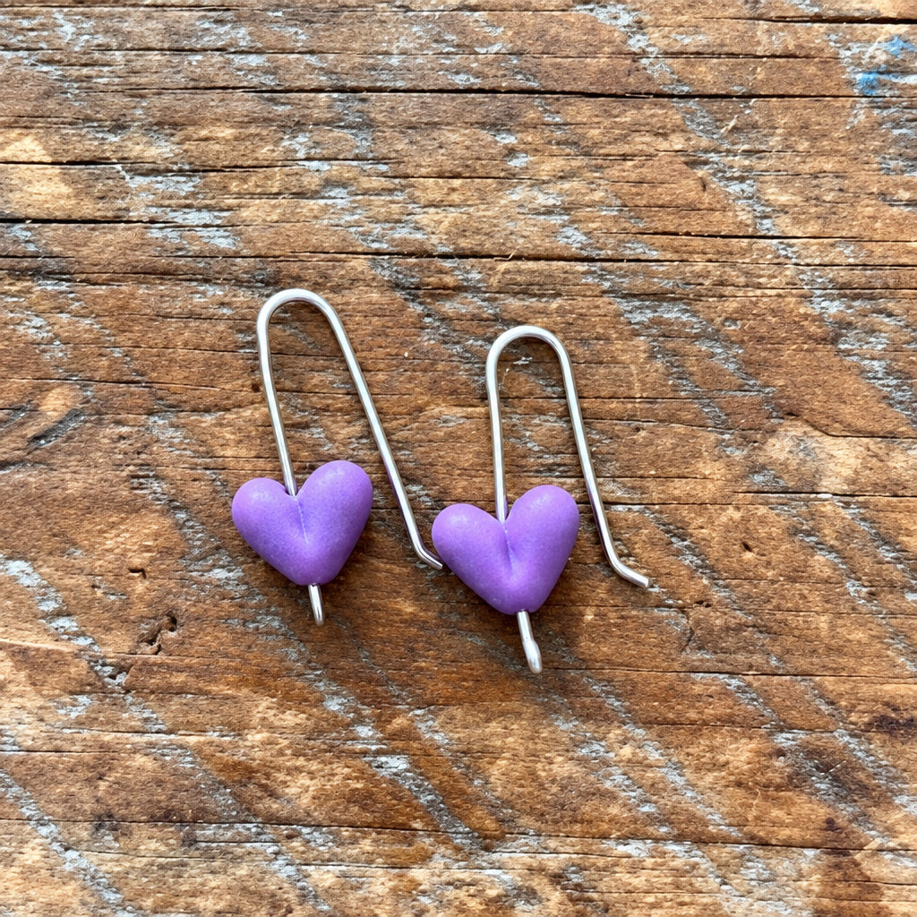 A pair of silver fishhook earrings with small, smooth purple heart-shaped beads, displayed on a rustic wooden surface.