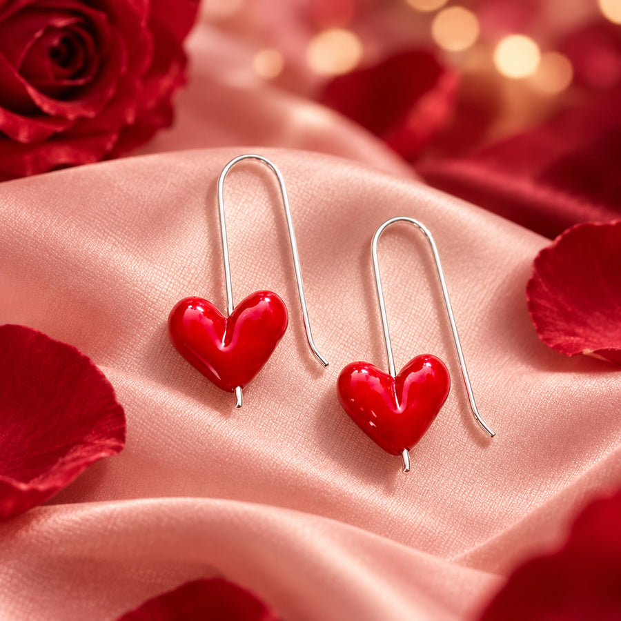 A pair of silver-tone drop earrings with glossy red heart-shaped charms, displayed on a textured off-white fabric background with a brand watermark in the top left corner.