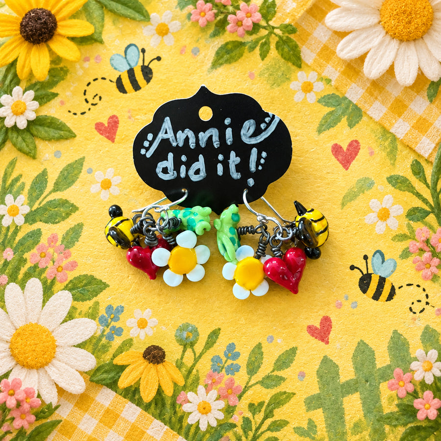 A pair of handmade polymer clay earrings featuring daisy flowers, red berries, green leaves, and bumblebees, hanging from a black display card that reads 'Annie did it!' on a rustic wooden background.