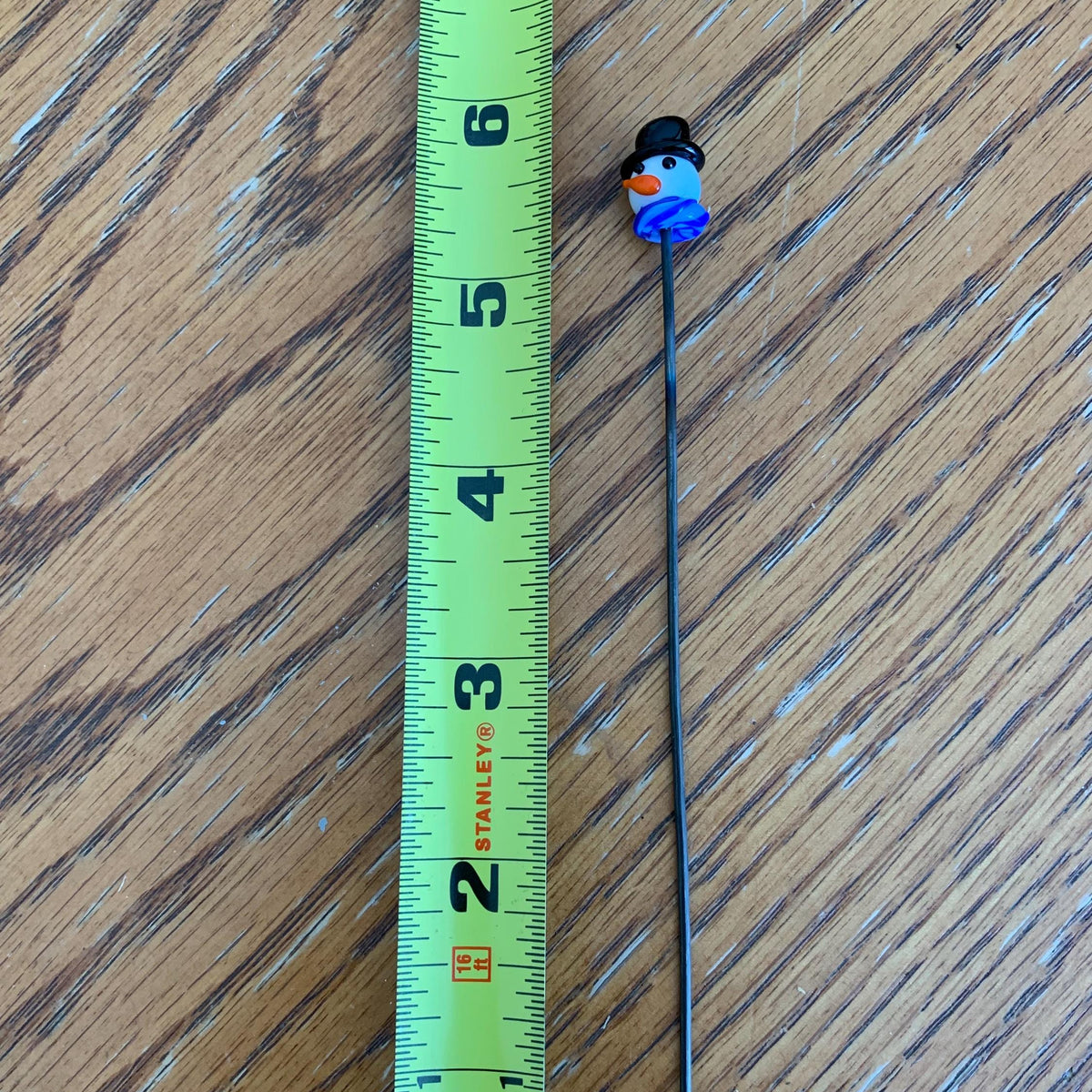 A long black pin or stick with a small snowman charm at the top is shown next to a green Stanley tape measure on a wooden surface. The snowman has orange eyes and a blue scarf.