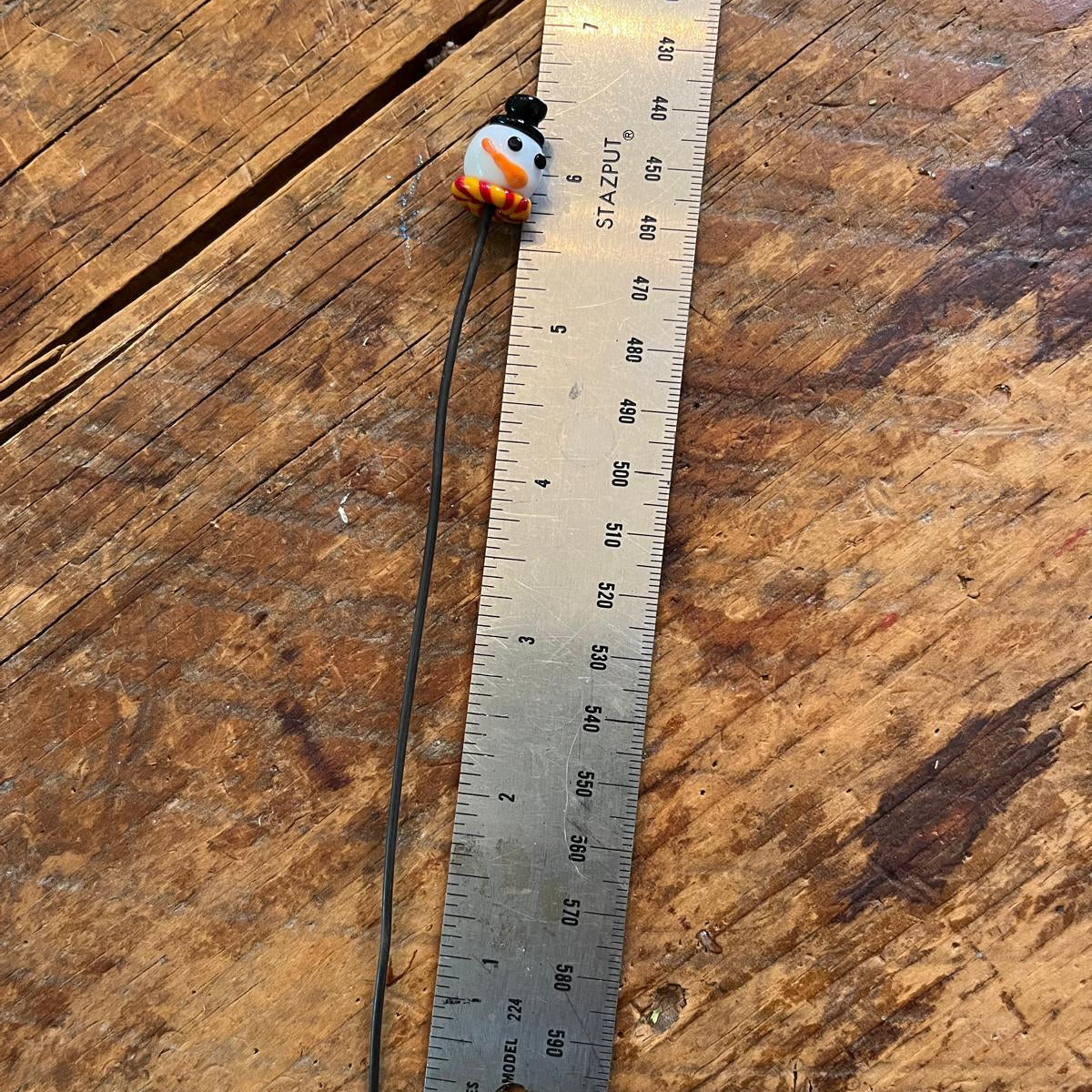 A single bead with a black wire tail, featuring a white and orange patterned top, shown next to a metal ruler on a rustic wooden surface.