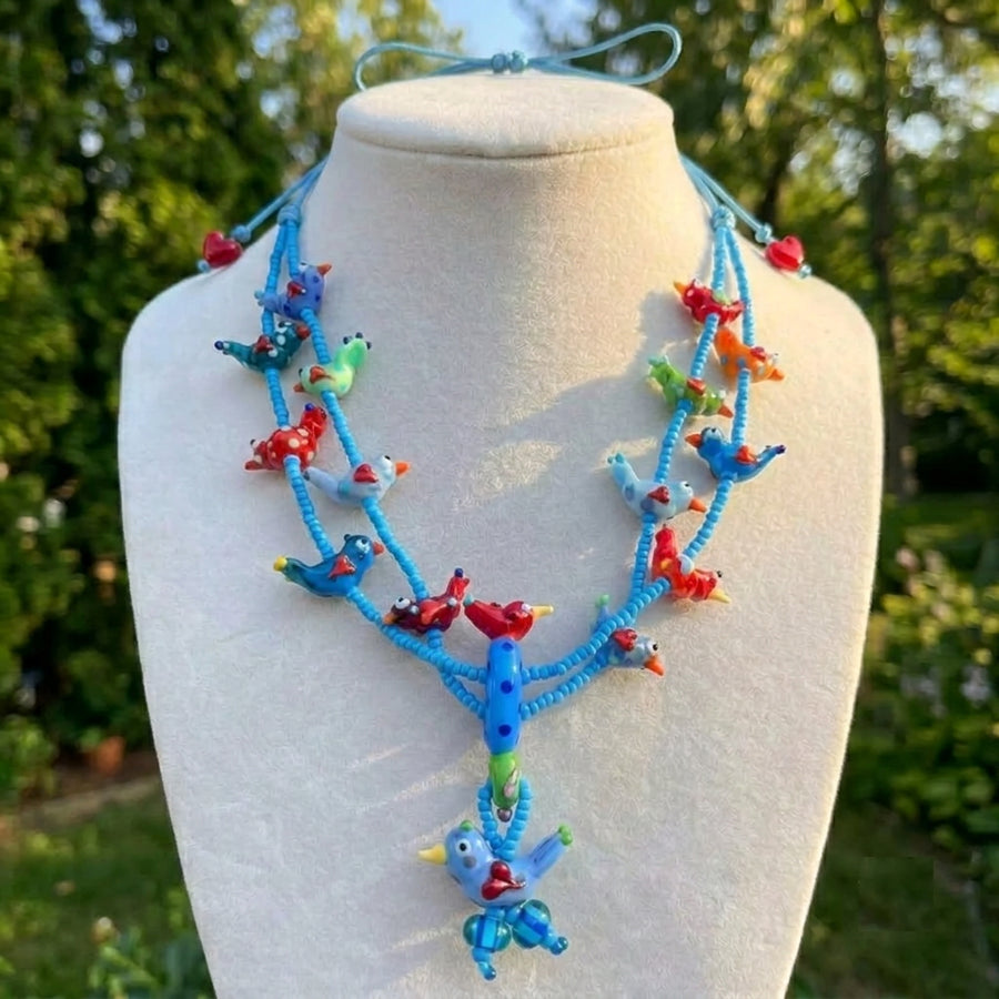 A multi-strand blue cord necklace with colorful bird charms, displayed on a cream-colored mannequin bust outdoors with a blurred green garden background. A text overlay in the top left reads 'Necklace Heart Wawa Collection - Blue'.