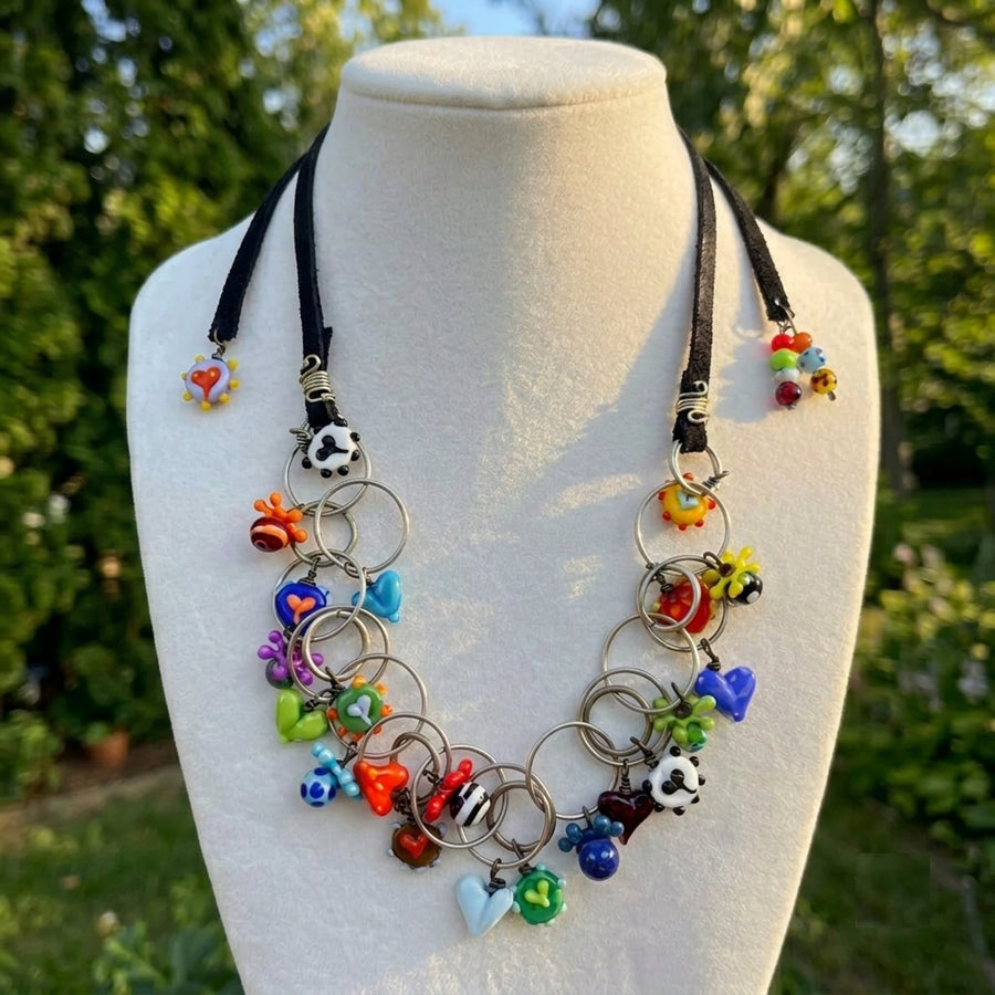 A colorful charm necklace displayed on a white mannequin bust outdoors. The necklace features black cords, silver-toned rings, and multiple small glass charms in various colors including blue, red, green, yellow, and white.