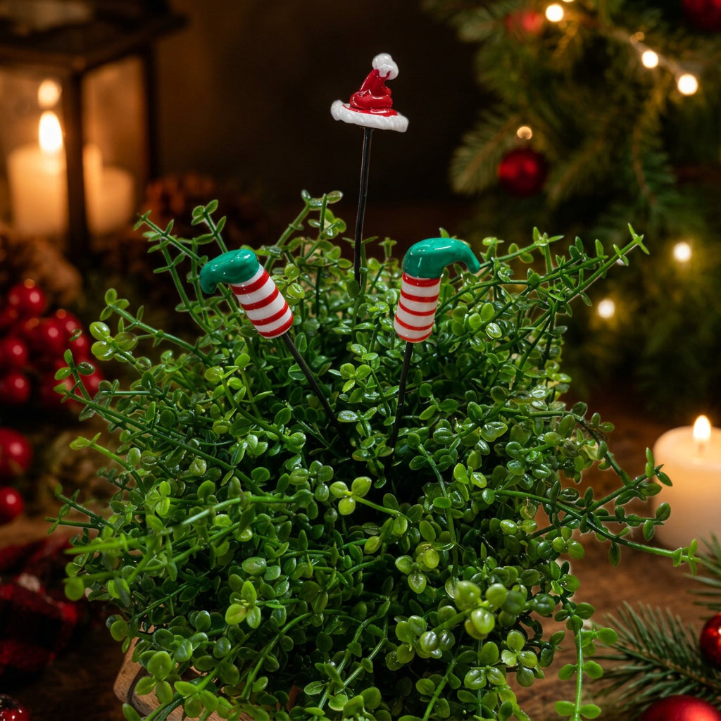 A festive Christmas plant stake decoration set featuring two green and red striped candy cane stakes and one Santa hat stake, displayed in a small green faux plant arrangement against a warm, blurred Christmas tree background.