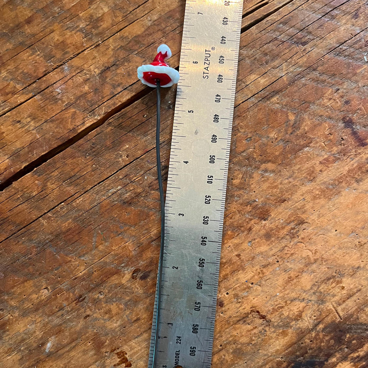 A miniature Santa hat accessory with a black wire stem, shown next to a metal ruler on a rustic wooden surface to indicate its small scale.
