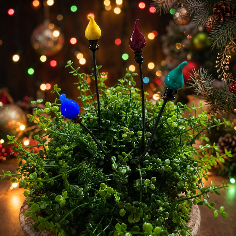 Four Christmas garden light plant sticks with colorful bird-shaped bulbs in yellow, red, blue, and teal, shown lit and planted in a green leafy pot against a dark garden background. The 'Plant Sticks' brand logo is visible in the top left.