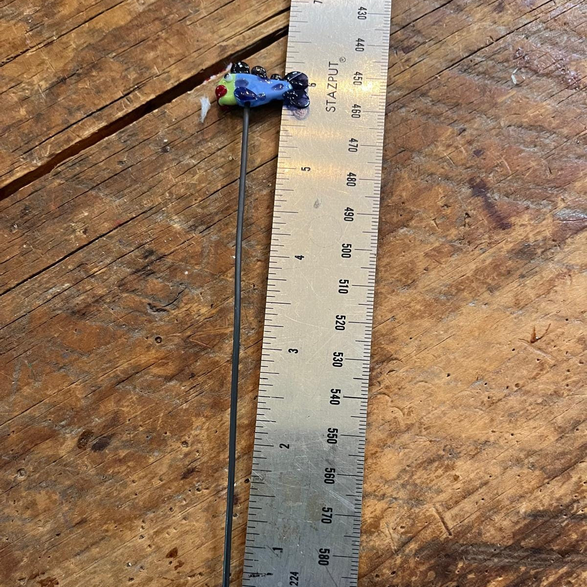 A very small blue and green insect-like specimen attached to a long thin needle, placed next to a metal ruler on a worn wooden tabletop.