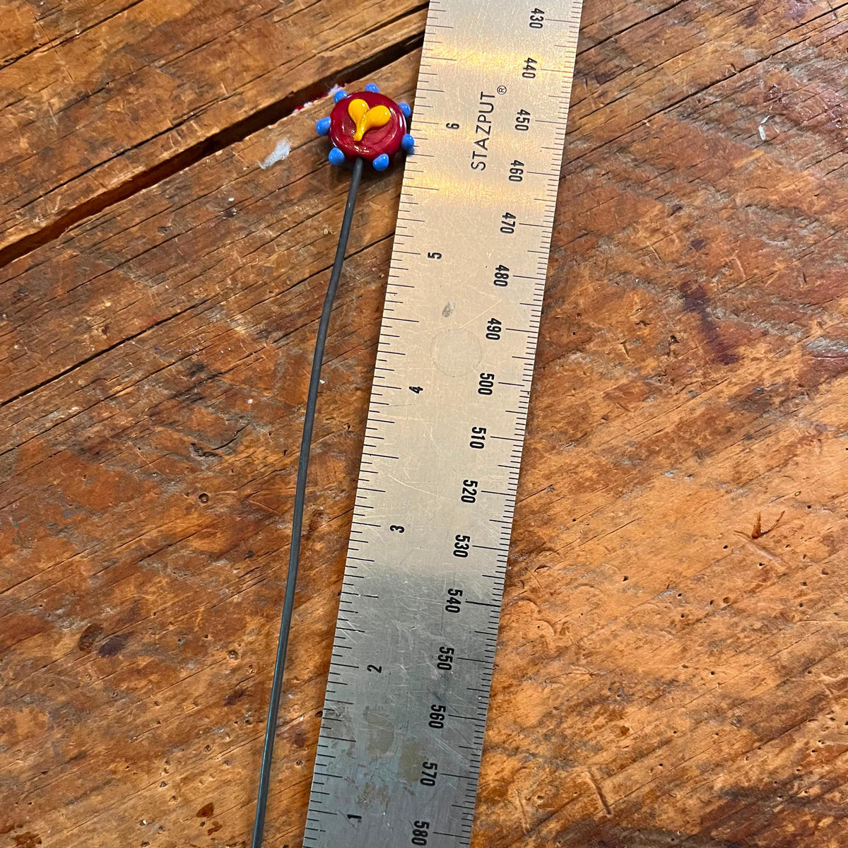 A long, thin black rod or stick with a small round red, yellow, and blue decorative end, placed next to a metal ruler on a rustic wooden surface.