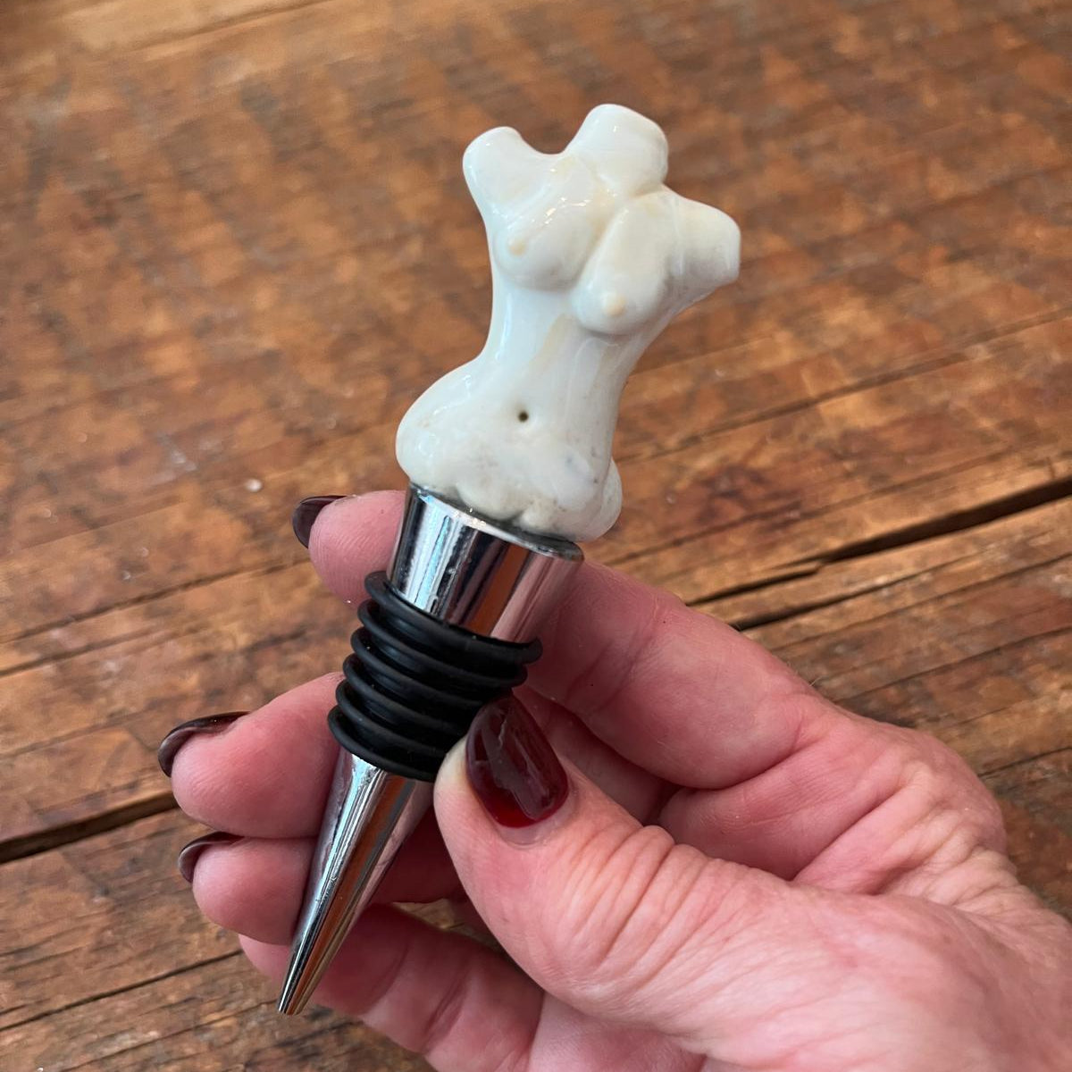A hand with dark red nail polish holds a wine bottle stopper featuring a bone-shaped white top and a black rubber sleeve on a silver metal body, set against a rustic wooden background.