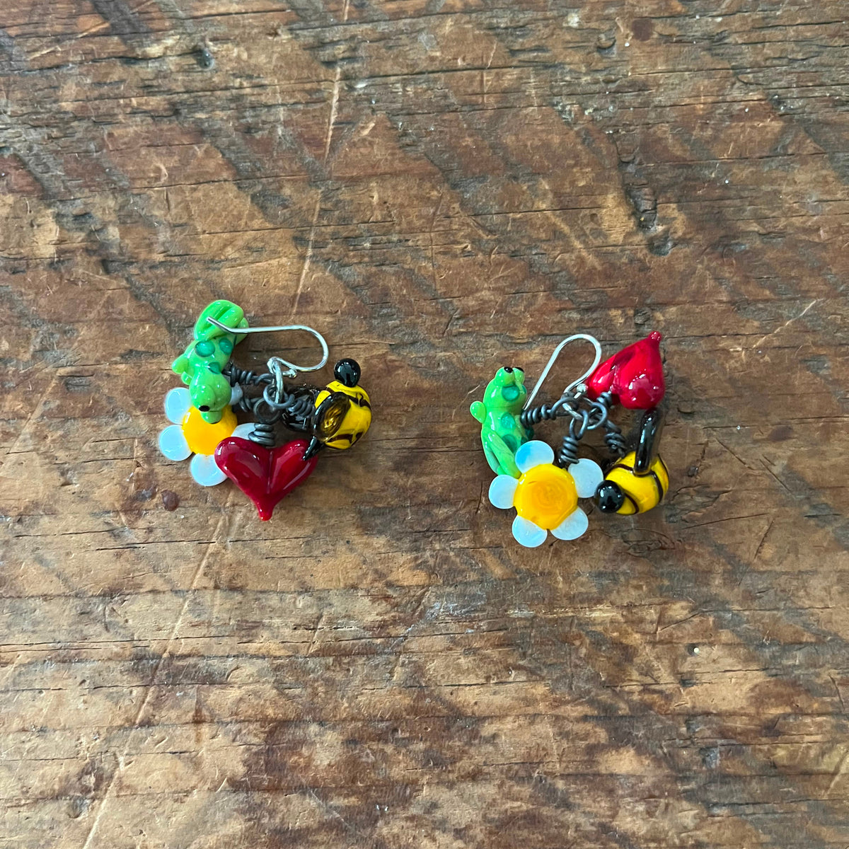 A pair of handmade, colorful earrings featuring bumblebees, flowers, and a red heart, displayed on a rustic wooden surface.
